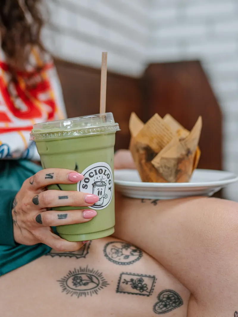 A guest holding an iced matcha in a Sociology cup, with pink nails and Sproey on the label
